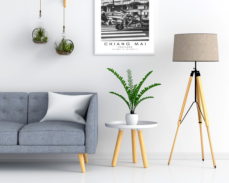 May include: A living room interior with a gray sofa, a white pillow, a potted plant, a small round wooden table, a tripod floor lamp with a beige lampshade, and a framed black and white photograph of a street scene in Chiang Mai, Thailand.