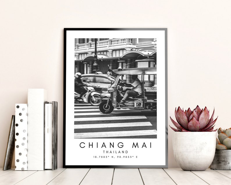 May include: Black and white photograph of a tuk-tuk driving down a street in Chiang Mai, Thailand. The tuk-tuk is a three-wheeled vehicle with a canopy and a driver in the front. The street is lined with buildings and there is a car in the background.