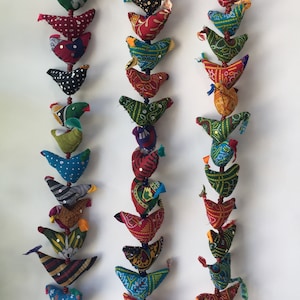 May include: Three strands of colourful fabric bird ornaments hanging from a white wall. Each strand has a bell at the bottom. The birds are made of patchwork fabric in various colours and patterns, including red, green, blue, yellow, and orange.
