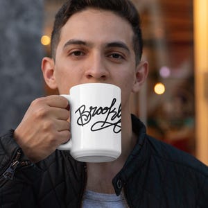 May include: White ceramic mug with the word "Brooklyn" in black cursive script. The mug is held up to the mouth. The person is wearing a black jacket and a gray shirt.