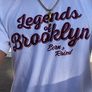 May include: White t-shirt with the text "Legends of Brooklyn Born & Raised" in maroon script. A gold chain with a pendant hangs around the neck. The pendant is a gold horn with a crown.