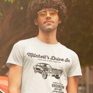 May include: A white t-shirt with a black and white illustration of a classic car. The text "Mitchell's Drive In" is printed in a retro font, along with the tagline "Your car is our counter". The text "At 86th St. & 7th Ave. Brooklyn New York stoop society" is printed below the car.