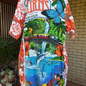 May include: A short-sleeve shirt featuring a vibrant graphic print of Cairns, Australia, including a large blue butterfly, a train on a bridge, a waterfall, a marlin, and tropical fish. The sleeves and sides have an orange floral pattern.