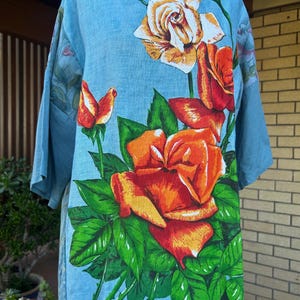 May include: A light blue short-sleeved top featuring a large, vibrant floral print of orange and red roses with green leaves. The design includes several blooming roses and buds scattered across the front, set against a solid light blue background.