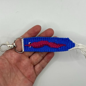 May include: A blue woven keychain with a pink worm design. The keychain has a silver clasp and a white tassel. The worm is a bright pink color and is centered on the blue background.