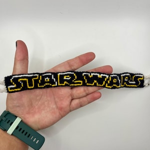 Op de afbeelding: Een zwart en geel geborduurd armband met de tekst "STAR WARS" in wit en geel. De armband wordt in een hand gehouden, met een groen horloge zichtbaar op de pols.