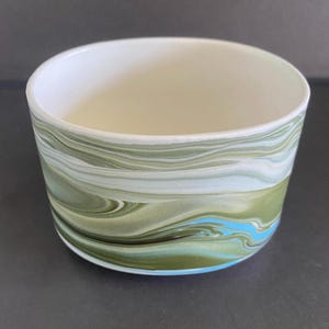 May include: A cylindrical ceramic bowl featuring a marbled pattern on the exterior with swirling shades of olive green, sage, white, and bright turquoise. The interior of the bowl is a solid, smooth off-white colour with a clean, rounded rim edge.