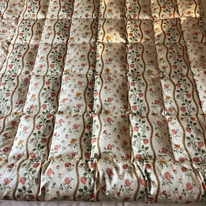 May include: A quilted comforter featuring a cream-coloured background with a repeating pattern of vertical wavy brown lines and small floral clusters in shades of red, orange, and green. The fabric is stitched into a grid of rectangular sections.