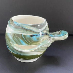 May include: A ceramic mug with a rounded body and a side handle, featuring a marbled glaze pattern in shades of blue, olive green, white, and grey. The interior is smooth and white, while the exterior displays a swirling, multi-coloured finish.
