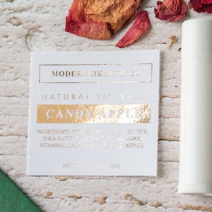 May include: A white tube of natural lip balm with a gold label that reads "MODERN HEALTH CO" and "CANDY APPLE". The label also lists the ingredients: cocoa butter, shea butter, coconut oil, beeswax, vitamin E, and crushed apples. The tube is 0.15 oz (4.2 g).