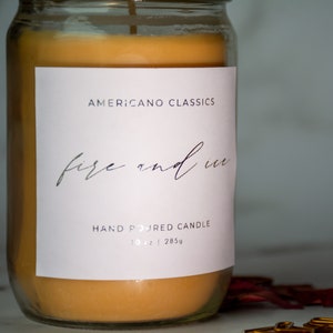 May include: A glass jar candle with a white label that reads "Americano Classics" and "fire and ice" in cursive script. The label also says "Hand Poured Candle" and "10 oz | 285g". The candle is a light orange color.