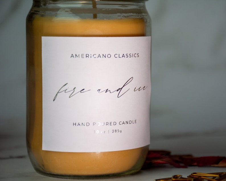 May include: A glass jar candle with a white label that reads "Americano Classics" and "fire and ice" in cursive script. The label also says "Hand Poured Candle" and "10 oz | 285g". The candle is a light orange color.