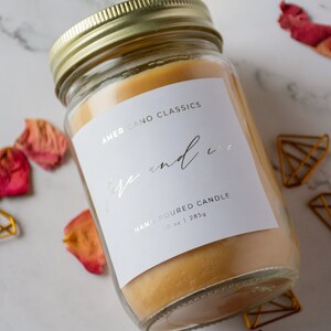 May include: A clear glass jar candle with a white label that reads "Americano Classics Fire and Ice Hand Poured Candle 10 oz | 285g". The candle is a light yellow color and is sitting on a white marble surface.