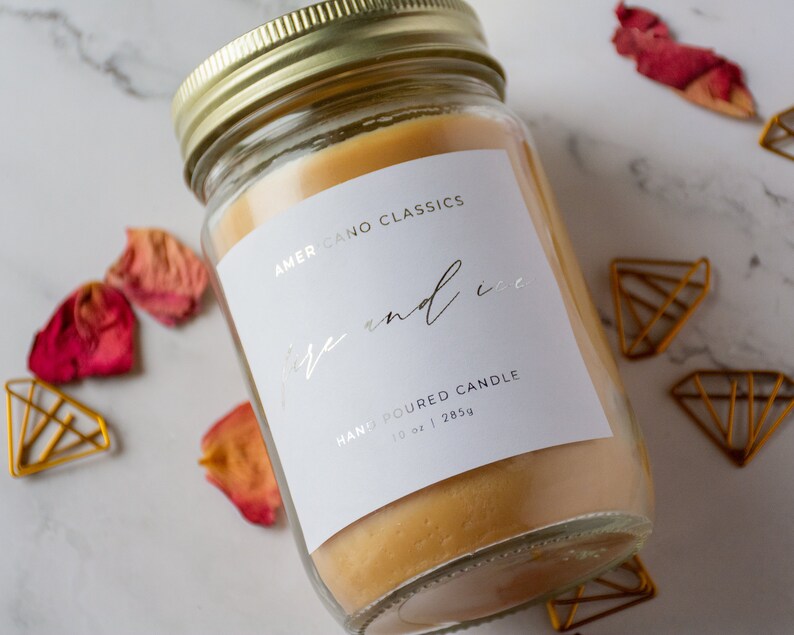 May include: A clear glass jar candle with a white label that reads "Americano Classics Fire and Ice Hand Poured Candle 10 oz | 285g". The candle is a light yellow color and is sitting on a white marble surface.