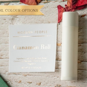 Peut inclure: Une étiquette blanche avec un lettrage en feuille d'or qui indique "MODERN PEOPLE" et "Cinnamon Roll". L'étiquette indique les ingrédients et le poids net de 4,2 g (0,15 oz). Un tube de baume à lèvres blanc est à côté de l'étiquette.