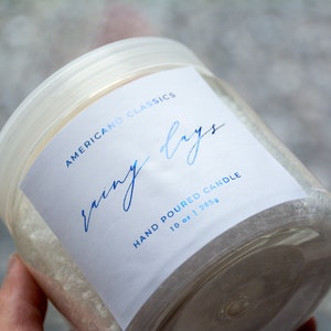 May include: A clear glass jar with a white label that reads "Americano Classics" and "Rainy Days" in blue script. The label also says "Hand Poured Candle" and "10 oz | 283g".