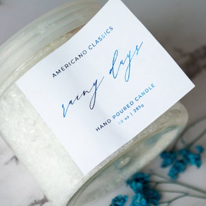 May include: A white glass jar with a white label that reads "Americano Classics Rainy Days Hand Poured Candle 10 oz | 285g".