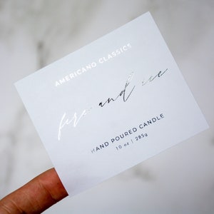 May include: White card with silver foil lettering. The text reads "Americano Classics fire and ice" and "Hand Poured Candle 10 oz | 285g".
