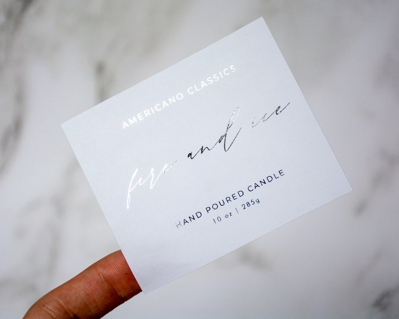 May include: White card with silver foil lettering. The text reads "Americano Classics fire and ice" and "Hand Poured Candle 10 oz | 285g".