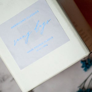 May include: White candle box with a blue label that says "Americano Classics" and "cozy days" in a handwritten font. The label also says "Hand Poured Candle 10 oz 140 hr."