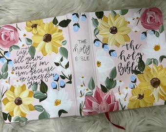 May include: A pink floral design covers the front and back of a closed Bible. The text "The Holy Bible" is written in black cursive on the front cover. The text "cast all your anxiety on Him because He cares for you" is written in black cursive on the back cover.