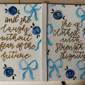 Może przedstawiać: Dwupanelowe płótno z ręcznie malowanym tekstem i kwiatowymi akcentami. Lewy panel głosi "i śmieje się bez strachu przed przyszłością" w złotym skrypcie. Prawy panel mówi "jest odziana w siłę i godność". Niebieskie kokardy i kwiaty zdobią białe tło.