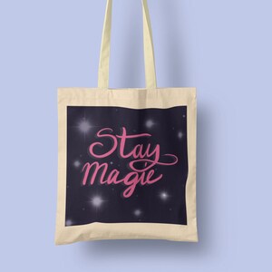 May include: A beige canvas tote bag with a black background and white stars. The text "Stay Magic" is written in pink cursive lettering.