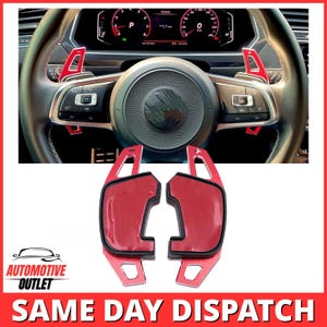 May include: A pair of red metal steering wheel paddle shifters with black trim and adhesive backing, shown both as individual components and installed on a black leather steering wheel with silver buttons and a digital dashboard display.