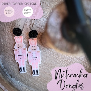 May include: A pair of pink and white nutcracker dangle earrings with black glitter accents. The earrings are on a wooden surface with the text "Nutcracker Dangles" in pink.