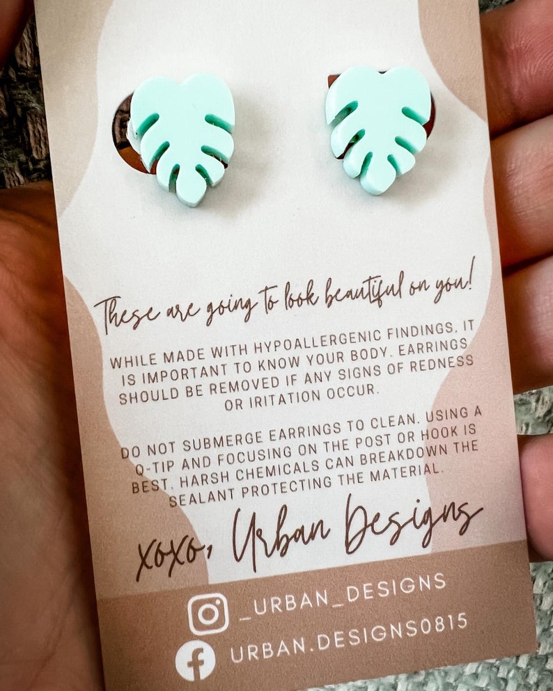 May include: A pair of light blue monstera leaf earrings on a white card with the text "These are going to look beautiful on you!" and "xoxo, Urban Designs" in brown text. The card also includes social media handles for Urban Designs.