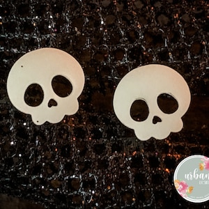 May include: Two white skull-shaped cutouts on a black sparkly background. The skulls have black eye sockets and a black nose.