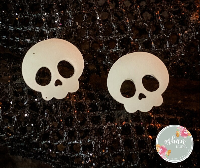 May include: Two white skull-shaped cutouts on a black sparkly background. The skulls have black eye sockets and a black nose.