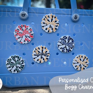 May include: A light blue tote bag with personalized cheer pom-pom charms. The charms are decorated with different colors and names, including "Tally", "Leigha", "Farrell", and "Cameron".