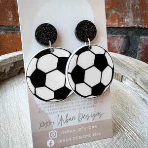 May include: A pair of black and white soccer ball earrings with black glitter accents. The earrings are on a white card with the text "xoxo, Urban Designs" and social media handles.