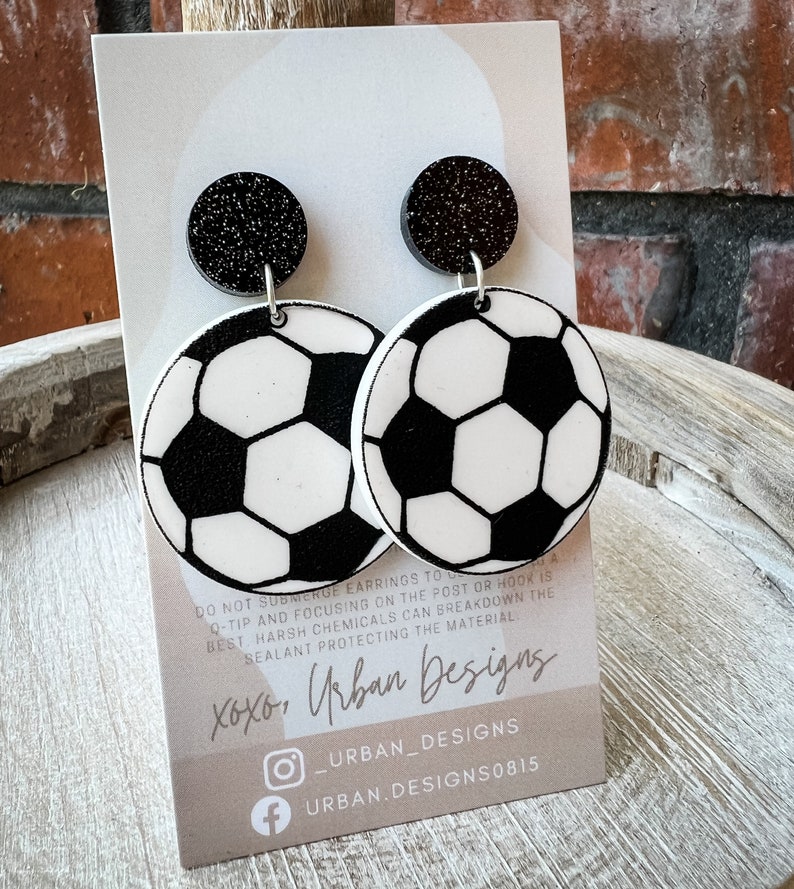 May include: A pair of black and white soccer ball earrings with black glitter accents. The earrings are on a white card with the text "xoxo, Urban Designs" and social media handles.