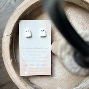 Peut inclure: Une paire de boucles d'oreilles en forme de fantôme blanc avec des yeux noirs et un petit sourire. Les boucles d'oreilles sont présentées sur une carte avec le texte "These are going to look beautiful on you!" et des informations sur les boucles d'oreilles et le vendeur, "Xoxo, Urban Designs".