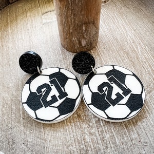 May include: A pair of black and white soccer ball earrings with the number 21 printed on each ball. The earrings have black glitter accents.