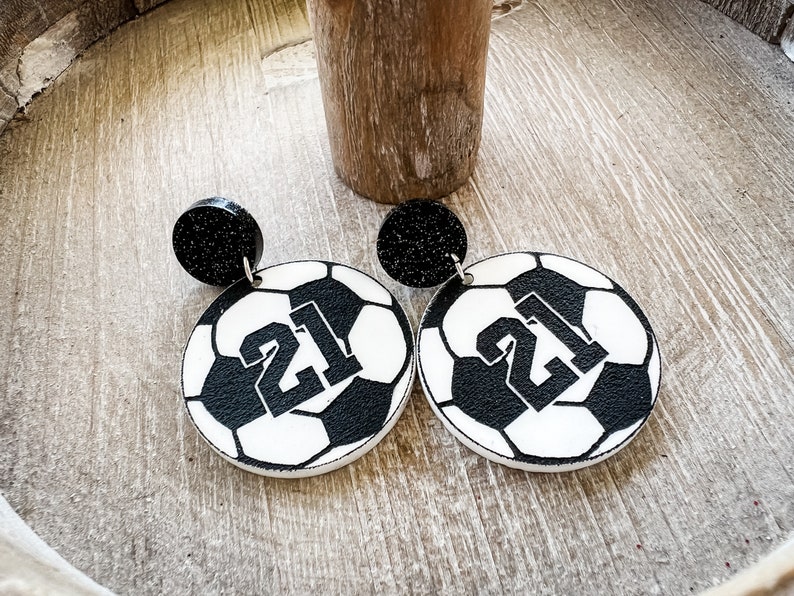 May include: A pair of black and white soccer ball earrings with the number 21 printed on each ball. The earrings have black glitter accents.