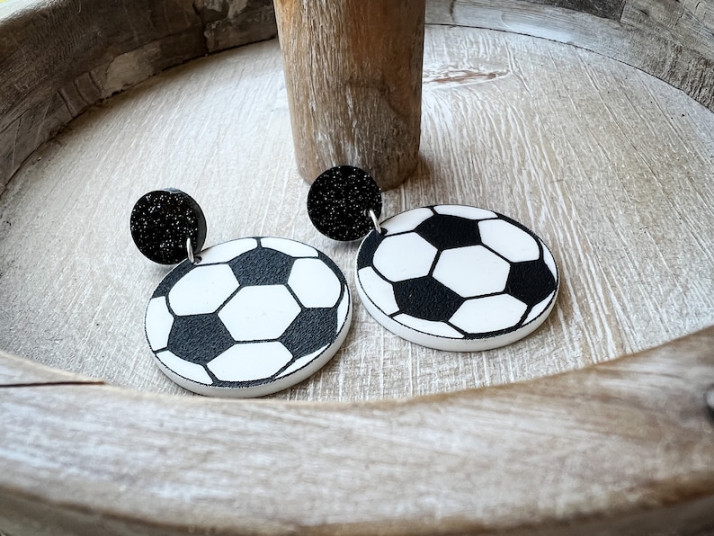 May include: A pair of black and white soccer ball earrings with black glitter accents. The earrings are made of polymer clay and are lightweight and easy to wear.