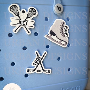 May include: Three personalized Bogg charms featuring a pair of lacrosse sticks with the name "JOEY", a pair of hockey sticks with the name "TROY", and a figure skate with the name "Airyn".