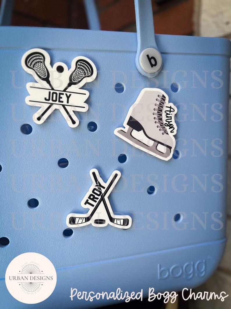 May include: Three personalized Bogg charms featuring a pair of lacrosse sticks with the name "JOEY", a pair of hockey sticks with the name "TROY", and a figure skate with the name "Airyn".