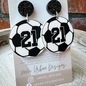 May include: A pair of black and white soccer ball earrings with the number 21 on each ball. The earrings have black glitter tops and silver hooks. The earrings are displayed on a card with the text "xoxo, Urban Designs" and social media handles.