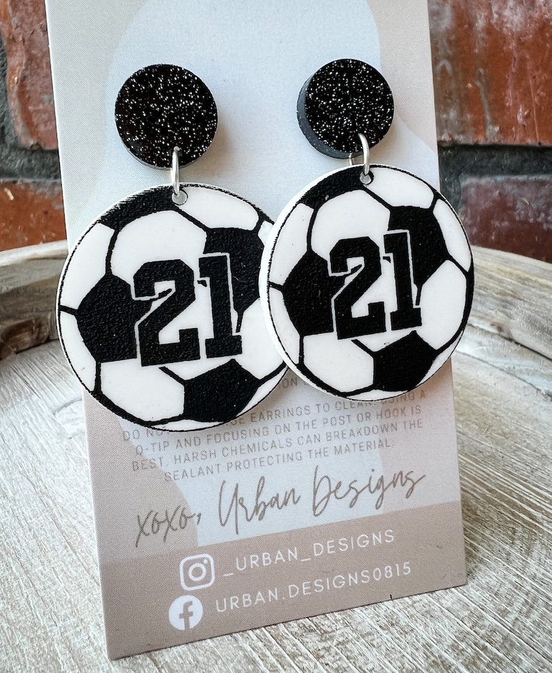 May include: A pair of black and white soccer ball earrings with the number 21 on each ball. The earrings have black glitter tops and silver hooks. The earrings are displayed on a card with the text "xoxo, Urban Designs" and social media handles.