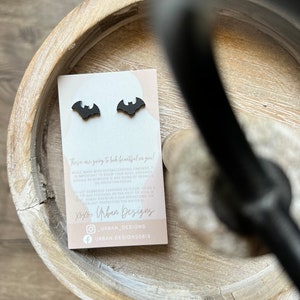 May include: A pair of black bat-shaped earrings on a pink card with the text "These are going to look beautiful on you!"  "While made with hypoallergenic findings, it is important to know your body. Earrings should be removed if any signs of redness or irritation occur." "Do not submerge earrings to clean. Using a tip and focusing on the post or hook is best. Harsh chemicals can breakdown the sealant protecting the material." "Xoxo, Urban Designs" "URBAN_DESIGNS" "URBAN.DESIGNS0815"