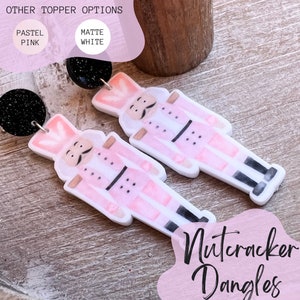 Peut inclure: Une paire de boucles d'oreilles pendantes en forme de casse-noisette rose et blanche avec des accents de paillettes noires. Les boucles d'oreilles sont en p&acirc;te polym&egrave;re et sont disponibles en rose pastel ou en blanc mat.