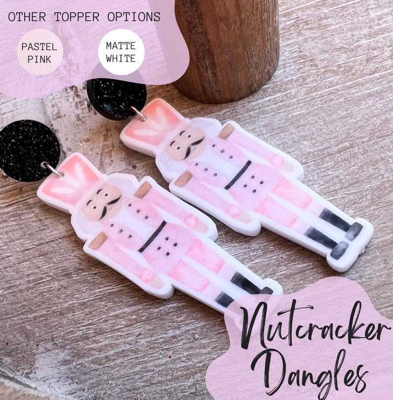 Peut inclure: Une paire de boucles d'oreilles pendantes en forme de casse-noisette rose et blanche avec des accents de paillettes noires. Les boucles d'oreilles sont en p&acirc;te polym&egrave;re et sont disponibles en rose pastel ou en blanc mat.