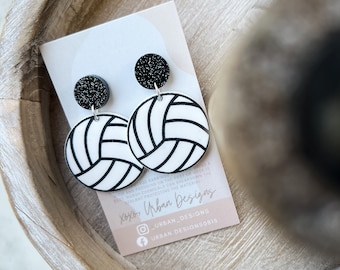 Könnte beinhalten: Ein Paar schwarze und weiße Volleyball-Ohrringe mit schwarzen Glitzerakzenten. Die Ohrringe befinden sich auf einer weißen Karte mit dem Text "xoxo, Urban Designs" und Social-Media-Handles.