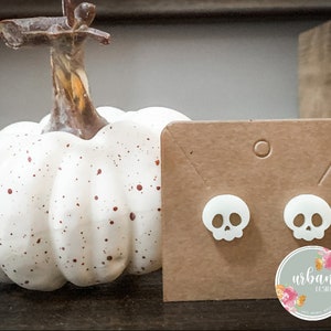 May include: A pair of white skull stud earrings on a brown card with the text "urban designs" and a floral design. The earrings are displayed next to a white pumpkin with brown spots.