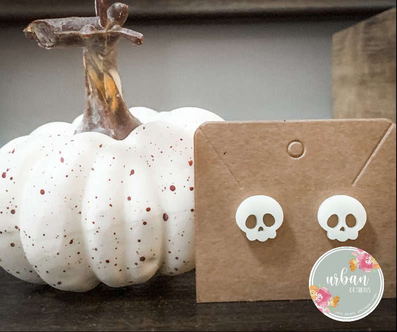 May include: A pair of white skull stud earrings on a brown card with the text "urban designs" and a floral design. The earrings are displayed next to a white pumpkin with brown spots.