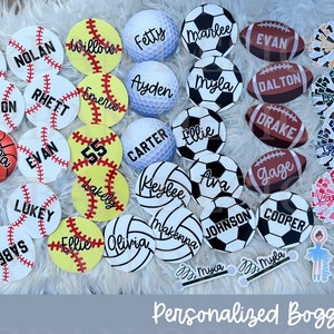 May include: A collection of personalized round sports-themed charms with various names and numbers. The charms are made of white, black, red, blue, and yellow with black lettering. The charms are arranged on a white fluffy surface. The text "Personalized Bogg Charms" is at the bottom of the image.